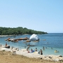 Strand mit Schwimmern und Hindernisparcours im Meer, bewaldete Küste im Hintergrund.