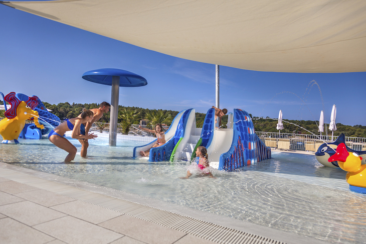 Kinder spielen auf Wasserrutschen in einem Pool mit Sonnensegel.