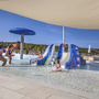 Kinder spielen auf Wasserrutschen in einem Pool mit Sonnensegel.