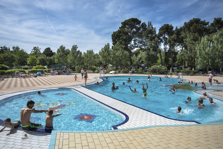 Menschen genießen einen sonnigen Tag am Pool mit blauen Himmel und Bäumen im Hintergrund.