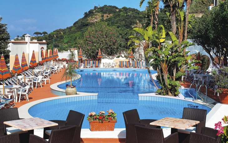 Hotelpool mit Liegestühlen und Sonnenschirmen, umgeben von Pflanzen und Bäumen.