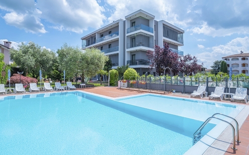 Hotel mit Pool und Liegestühlen bei Sonnenschein, Bäume und Gebäude im Hintergrund.