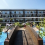 Hotel mit Holzbrücke und Pool, umgeben von Pflanzen bei sonnigem Himmel.