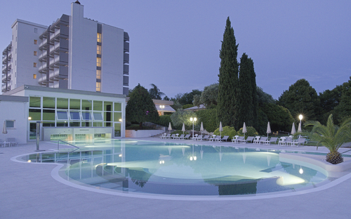 Nachtansicht eines beleuchteten Hotelpools mit Liegestühlen und Bäumen im Hintergrund.