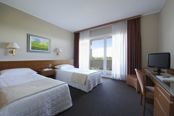Hotelzimmer mit zwei Einzelbetten, Schreibtisch und Balkon mit Vorhängen.