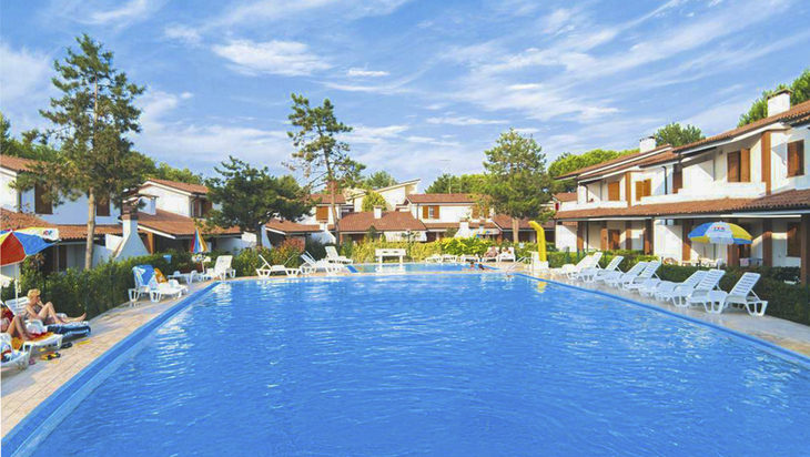 Ferienanlage mit großem Pool und Sonnenliegen, umgeben von Häusern und Bäumen.