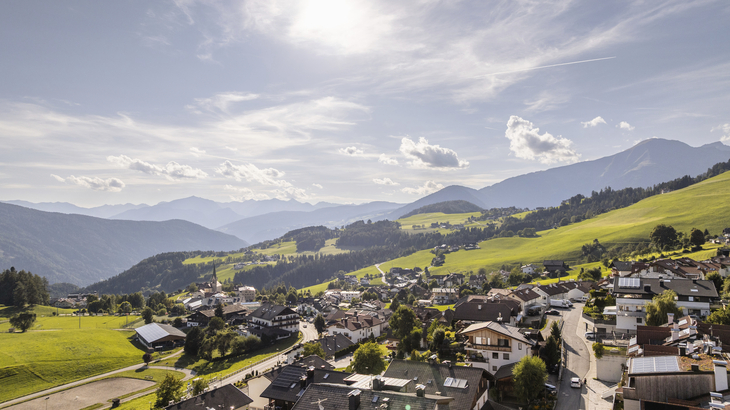 Bergdorf mit grünen Wiesen und Alpenlandschaft im Hintergrund bei sonnigem Wetter.