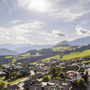 Bergdorf mit grünen Wiesen und Alpenlandschaft im Hintergrund bei sonnigem Wetter.