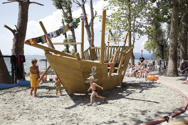Kinder spielen auf einem Spielplatz mit Holzschiff in Ufernähe.