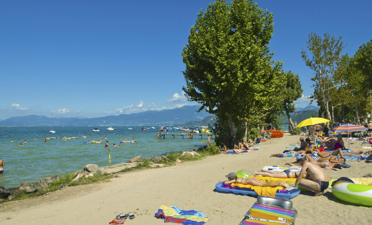 Strand am Gardasee mit Menschen beim Entspannen und Baden unter blauem Himmel.