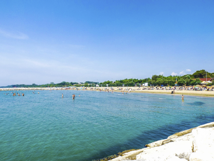 Strand mit Badegästen am Meer bei sonnigem Wetter, grüner Hintergrund.
