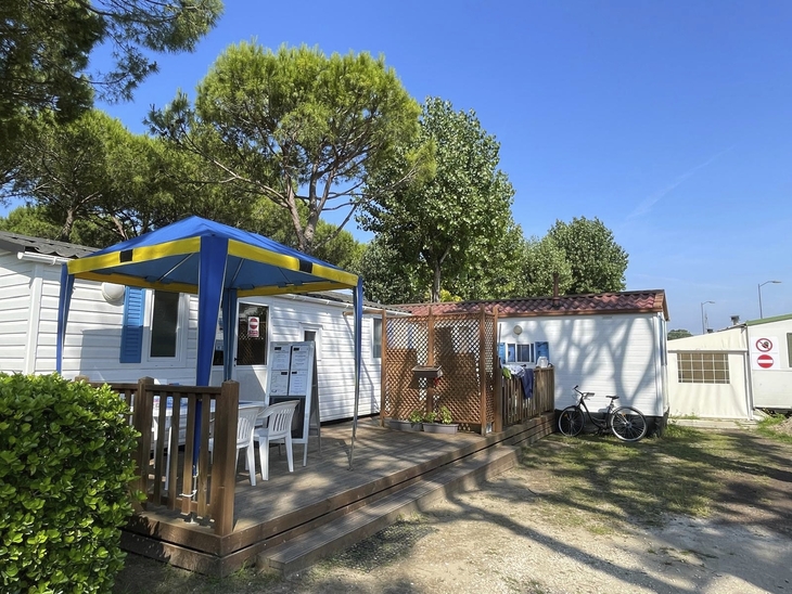 Campingplatz mit Mobilheim, Terrasse und Fahrrad, umgeben von Bäumen bei sonnigem Wetter.