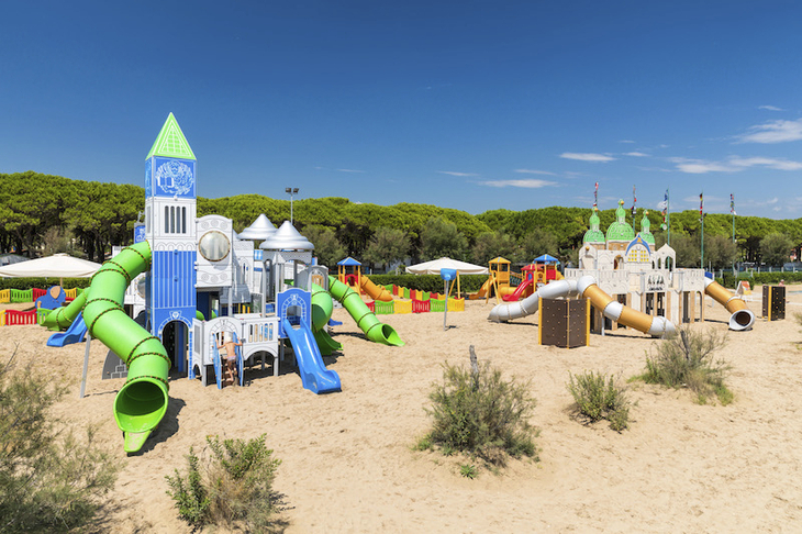 Spielplatz mit bunten Rutschen und Türmen auf Sand unter blauem Himmel.