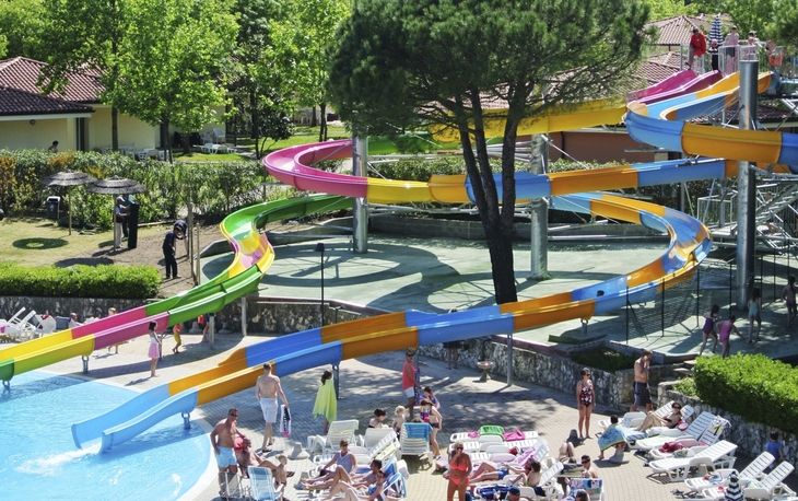 Bunte Wasserrutschen im Camping Bella Italia, umgeben von Sonnenliegen und Bäumen.