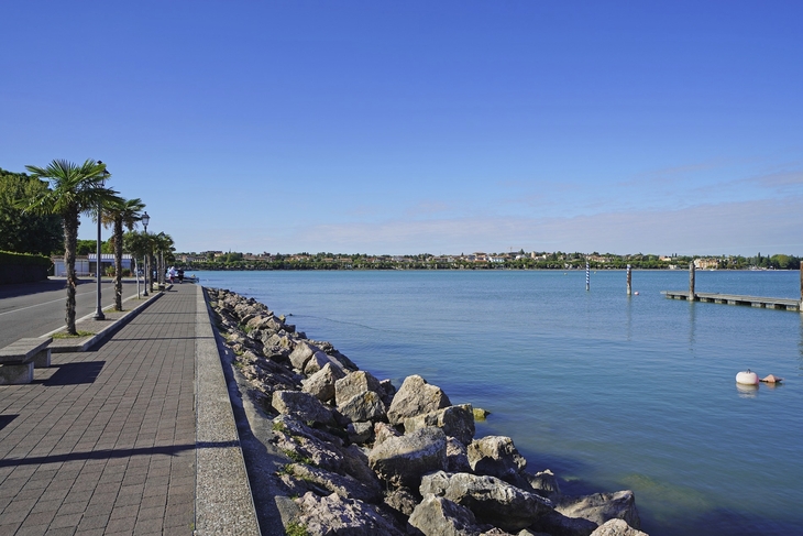 Promenade mit Palmen und Steinen am Ufer eines Sees unter blauem Himmel.