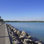 Promenade mit Palmen und Steinen am Ufer eines Sees unter blauem Himmel.