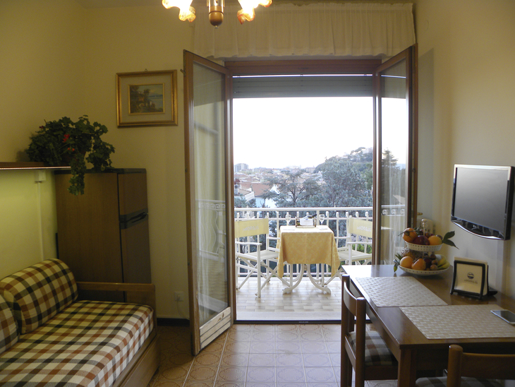 Wohnzimmer mit Balkon, Blick ins Freie, Tisch und Stühle, Sofa, Fernseher an der Wand.