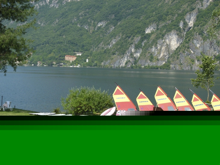 Sonnenliegen und bunte Segel am Ufer eines Sees vor Bergkulisse.