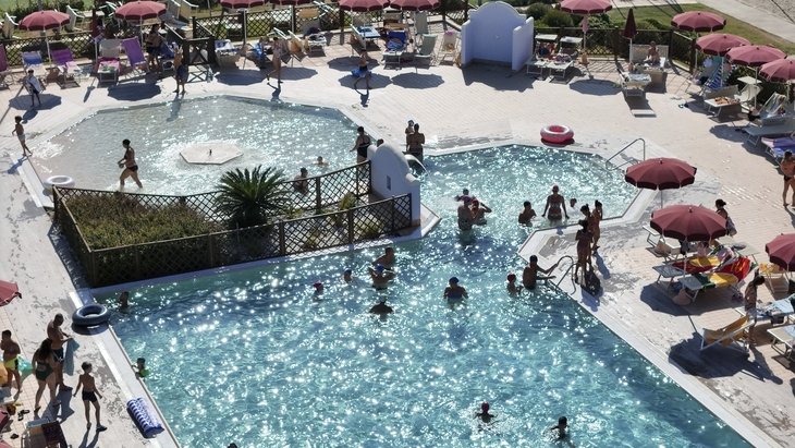 Menschen schwimmen und entspannen an einem sonnigen Pool mit roten Sonnenschirmen.