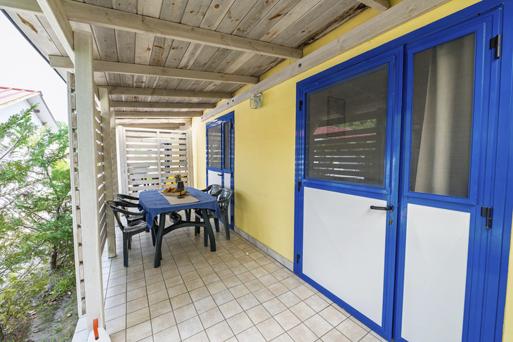 Überdachte Terrasse mit Tisch und Stühlen, gelbes Gebäude, blaue Tür und Fensterläden.