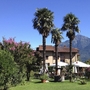 Garten mit Palmen und blühenden Büschen vor einem Haus, Berge im Hintergrund.