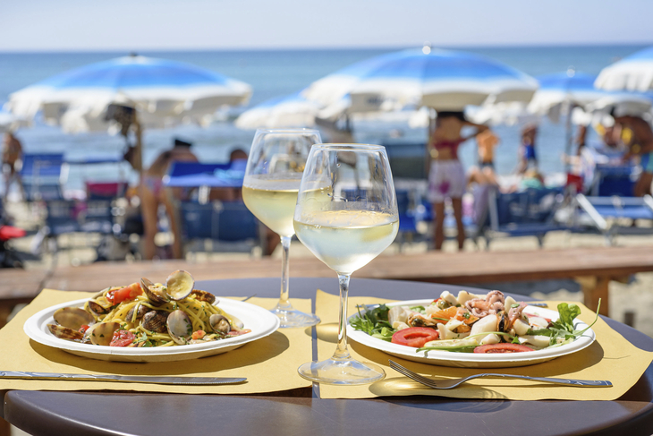 Zwei Teller mit Meeresfrüchtegerichten und Weißwein am Strandtisch.