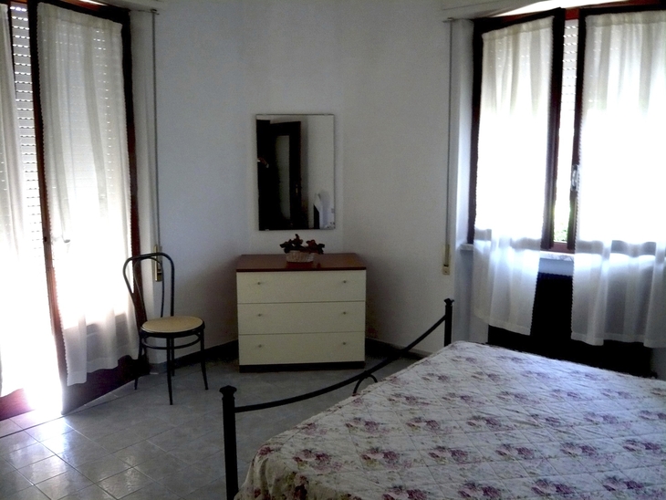 Ein Schlafzimmer mit Bett, Kommode, Stuhl und Vorhängen an den Fenstern.