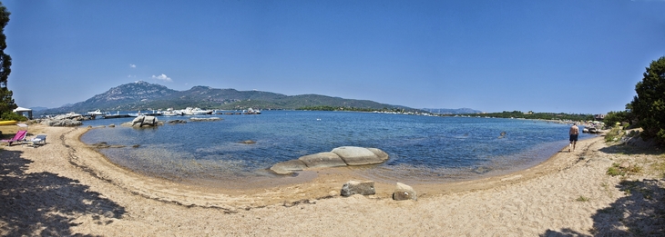 Panorama einer Bucht mit Sandstrand und klarem Wasser, umgeben von Bergen und Bäumen.