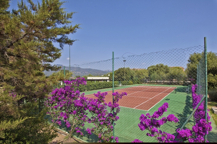 Tennisplatz mit violetten Blumen im Vordergrund, umgeben von Bäumen und blauem Himmel.