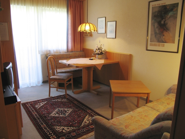 Gemütliches Wohnzimmer mit Tisch, Stühlen, Sofa und Teppich, hell durch Vorhangfenster.