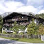 Landhaus mit Holzbalkon und Blumen, umgeben von Bäumen und Berglandschaft im Hintergrund.