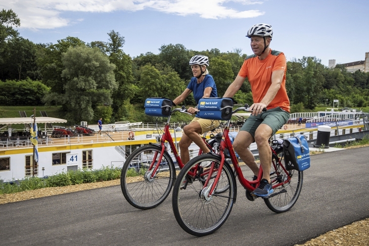 Zwei Personen fahren mit Fahrrädern auf einem Radweg entlang eines Flusses.