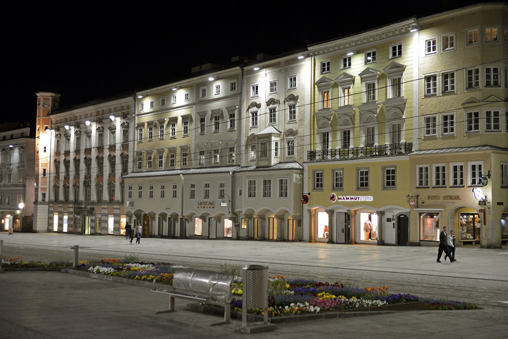 Historische Gebäude in Linz bei Nacht, beleuchtet mit Blumenbeeten im Vordergrund.