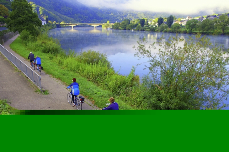 Radfahrer auf einem Weg entlang eines Flusses mit Brücke im Hintergrund.