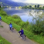 Radfahrer auf einem Weg entlang eines Flusses mit Brücke im Hintergrund.