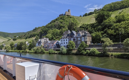 Schiff auf Fluss, Burgruine auf Hügel hinter Fachwerkhäusern, bewaldete Hänge im Hintergrund.