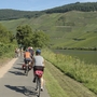 Radfahrer auf einem Weg entlang eines Flusses in einer grünen, hügeligen Landschaft.