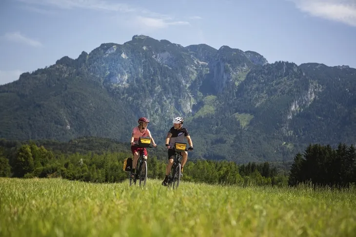 Zwei Radfahrer vor einer Bergkulisse auf einem grünen Feld.
