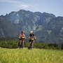 Zwei Radfahrer vor einer Bergkulisse auf einem grünen Feld.