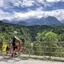 Radfahrer auf einer Straße mit Blick auf bewaldete Berge und bewölkten Himmel.