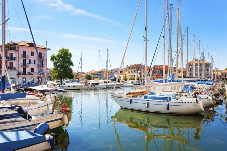 Boote im Hafen von Grado, umgeben von Gebäuden, unter blauem Himmel.