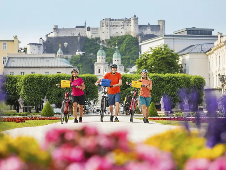 Drei Radfahrer im Mirabellgarten Salzburg mit Festung Hohensalzburg im Hintergrund.