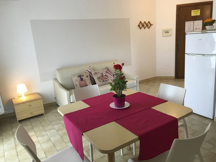 Esszimmer mit Tisch, roten Tischläufer, weißen Stühlen und Sofa im Hintergrund.