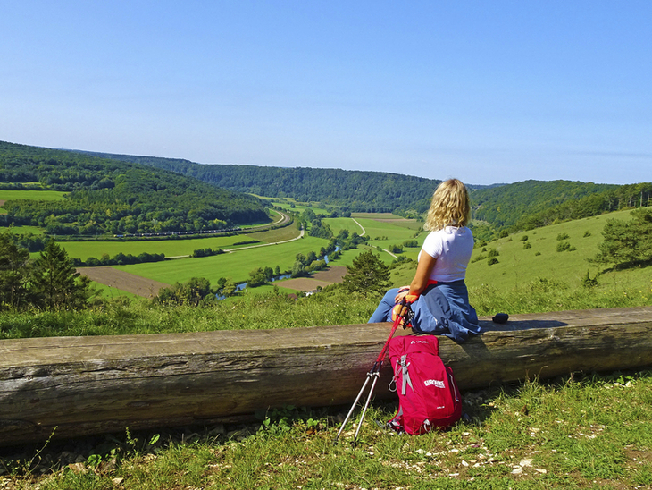 Frau sitzt auf Holzstamm am Altmühltal Panoramaweg mit Ausblick auf grüne Landschaft.