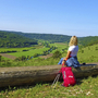 Frau sitzt auf Holzstamm am Altmühltal Panoramaweg mit Ausblick auf grüne Landschaft.