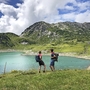 Zwei Wanderer am Formarinsee vor einer Bergkulisse bei sonnigem Wetter.