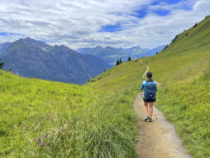 Person wandert entlang eines Pfades mit Bergblick und grünem Wiesenhang.