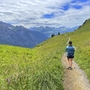 Person wandert entlang eines Pfades mit Bergblick und grünem Wiesenhang.