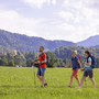 Drei Personen wandern auf einem grünen Feld vor einer Bergkulisse.