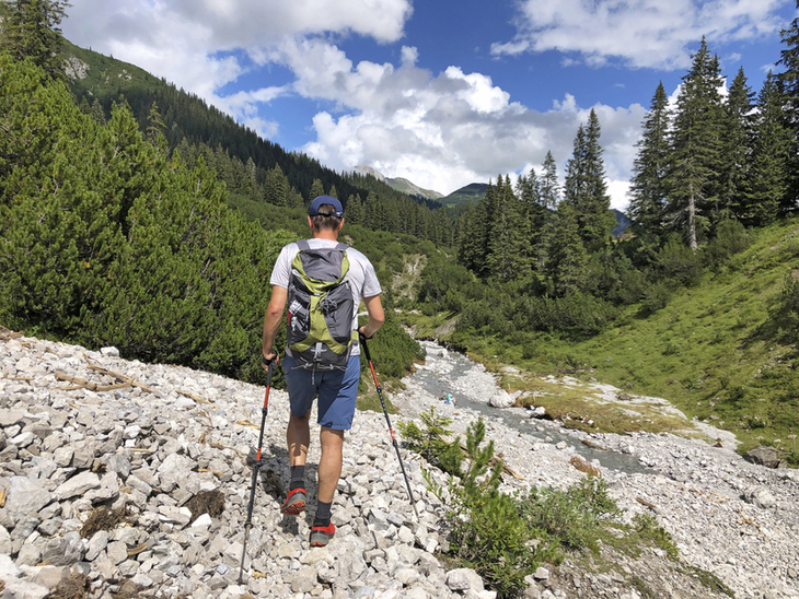 Wanderer mit Rucksack auf einem Bergweg in alpiner Landschaft mit Bäumen und Fluss.